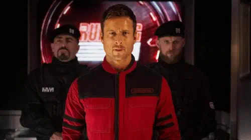 assets/glen-powell-is-escorted-by-two-men-in-the-film-the-running-man.jpeg