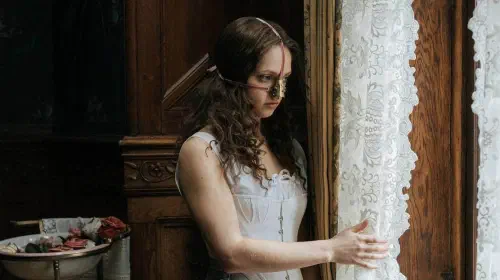 assets/a-woman-with-a-metallic-device-on-her-face-looks-out-a-window-in-the-ugly-stepsister.jpeg