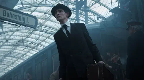 assets/kafka-in-a-suit-and-hat-leaning-out-from-a-crowd-in-franz.jpeg